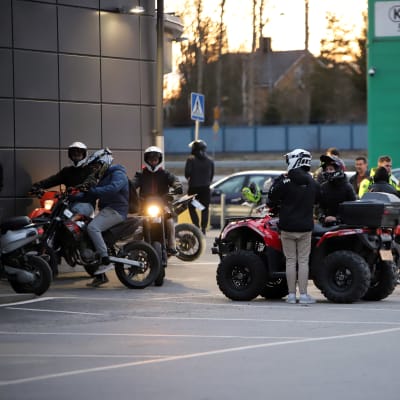 Ungdomar står med mopeder och fyrhjulingar på en parkering i kvällsljus.