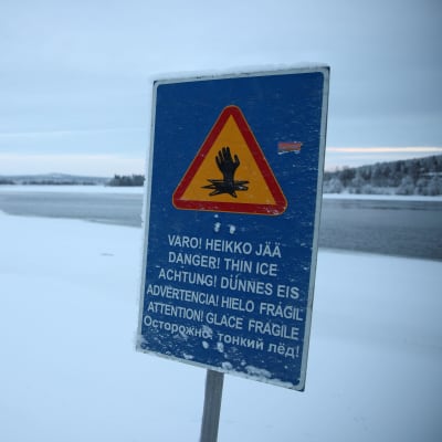 Kyltti Rovaniemellä, joka varoittaa heikosta jäistä. Taustalla osittain sula joki.