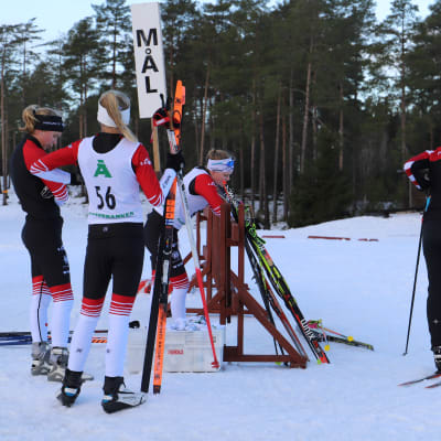 En grupp flickor på skidor står runt en stolpe där det står "mål".