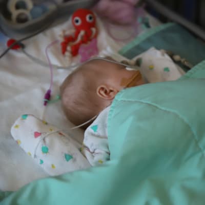 Pieni vauva nykkuu sairaalasängyssä vihreän peiton alla. Hengitysviikset on kiinnitetty kasvoille teipillä. Äiti silittää vauvaa peiton päältä.