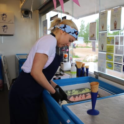 En ung kvinna med namnet Ursula Mantere arbetar som försäljare i en glasskiosk. I förgrunden syns en glassvåffla i en blå strut och Ursula håller på att skopa glass.
