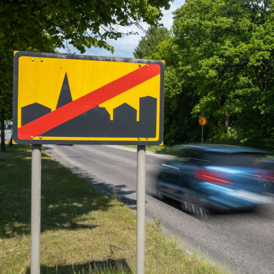 Ett trafikmärke som visar att tätort upphör syns i förgrunden, vid sidan av det två bilar som är fotade med rörelseoskärpa.