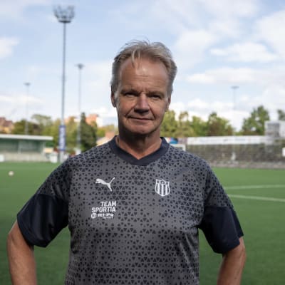 Juha Malinen seisoo jalkapallokentän laidalla ja katsoo kameraa kohti. Hänen yllään on paita, jossa on KTP-joukkueen logo.