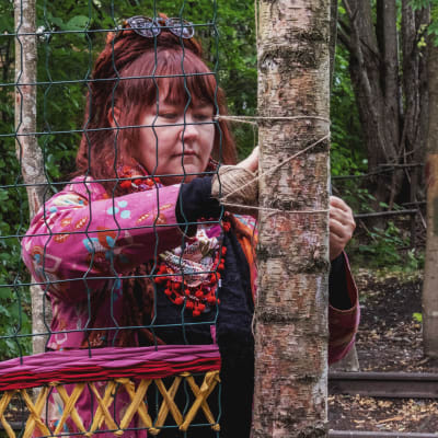 Kvinna i chockrosa och blommig rock och färggrann schal väver ett mönster med färggranna trådar på ett spjälverk som sitter fast mellan två träd.