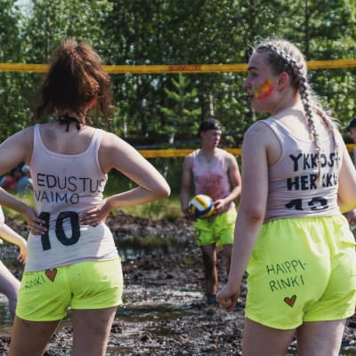 Unga kvinnor i gula shorts och vita linnen står till knäna insjunkna i en torvmosse och spelar kärrvolleyboll.