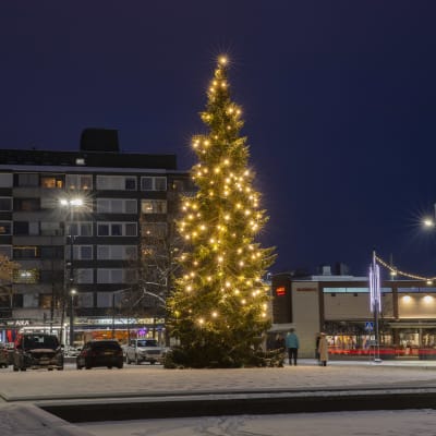 Jouluvaloilla koristeltu joulukuusi Kouvolan Kaupungintalon pihalla. Kuusen oikealla puolella Raimo Utriaisen veistos Polvesta polveen.