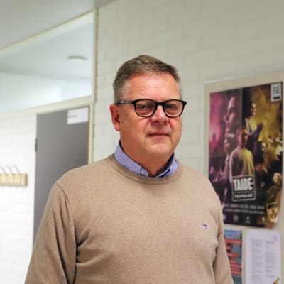 En man står i en skolkorridor. I bakgrunden syns vita stenväggar, en dörr till ett klassrum och klädkrokar där en jacka och en tygkasse hänger. Till höger om personen som är på fotot finns en anslagstavla med flera olika posters och blanketter.