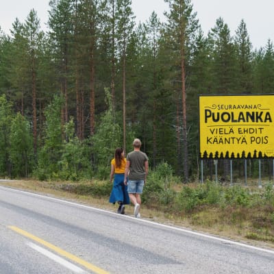 Nainen ja mies kävelevät maantien reunaa pitkin suuren, keltaisen kyltin ohi.