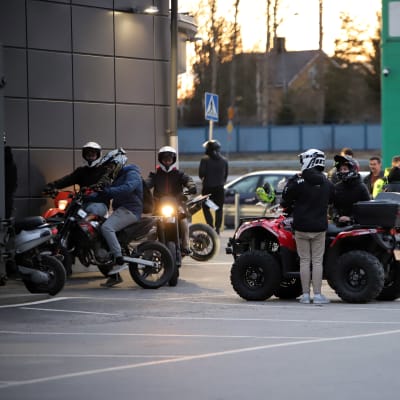 Ungdomar står med mopeder och fyrhjulingar på en parkering i kvällsljus.