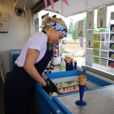 En ung kvinna med namnet Ursula Mantere arbetar som försäljare i en glasskiosk. I förgrunden syns en glassvåffla i en blå strut och Ursula håller på att skopa glass.