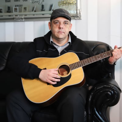 Tino Zitron istumassa sohvalla kitara käsissään.