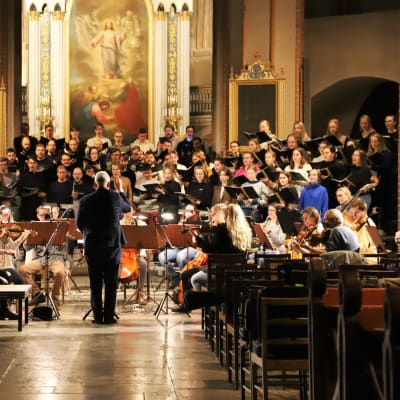 En man står med ryggen mot kameran och dirigerar en kör och en orkester i en kyrka. 