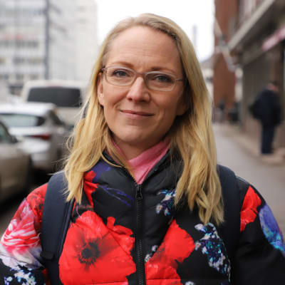En kvinna med allvarlig min står på en trottoar i Helsingfors och tittar in i kameran. 