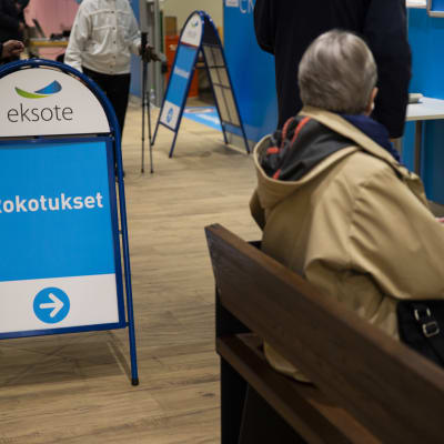 Eksoten rokotuspisteen kyltti sekä istuva nainen kauppakeskus IsoKristiinassa Lappeenrannassa.