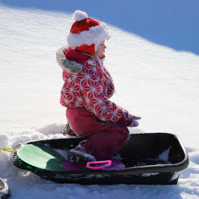 Liten flicka som sitter i en pulka i en snöig backe.
