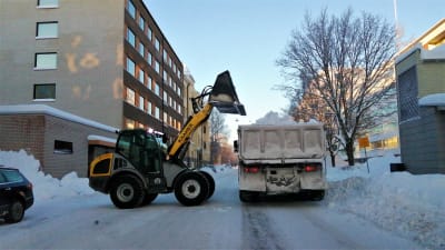 Snöplog lyfter upp lass med snö på flaket till en lastbil längs Skolhusgatan i Vasa.