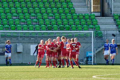 En klunga unga kvinnor i röd spelarklädsel gratulerar varandra glatt efter match. I bakgrunden ett fotbollsmål.