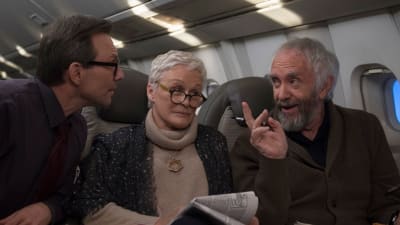 Joan (Glenn Close) sitter i ett flygplan och ser irriterad ut, omringad av Nathaniel (Christian Slater) och Joe (Jonathan Pryce) som pratar med varandra.
