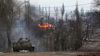 En rysk stridsvagn på en väg i Mariupol. I bakgrunden syns en byggnad i brand och kala träd.