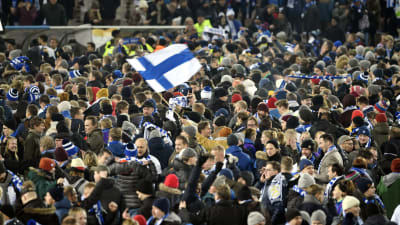 Ett myller av människor. Många med Finlands färger på mössa eller halsduk. En person håller i Finlands flagga.
