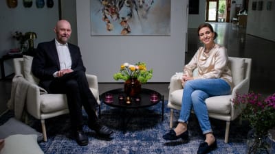 Esko Valtaoja och programledaren Ira Hammerman sitter i varsin vita fåtölj och småler till kameran. I bakgrunden hänger en tavla och mellan dem står ett litet bord med en vas med blommor i.