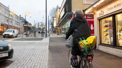 Kvinna cyklar på gatan, till höger affärslokal och till vänster motorhuven av en parkerad bil.