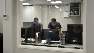 Två män tittar på datorskärmar i ett rum med ett glasfönster.
