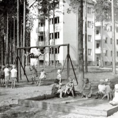 Mustavalkoisessa valokuvassa lapsia on kiikkumassa ja leikkimässä hiekkalaatikolla kerrostalon pihalla.