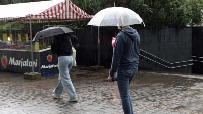 Två personer med paraply över huvudet går på en regnfylld gata i Helsingfors. De går förbi ett bärstånd.