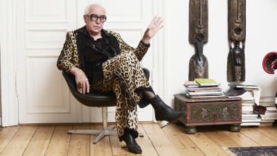 Ditlev Tamm sitter i en svart fåtölj, iklädd leopardmönstrad jacka och byxor, svart skjorta och svarta, högklackade skor. 