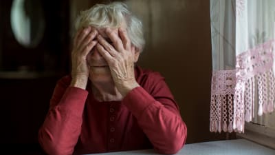 en äldre kvinna i röd blus håller händerna på ansiktet. Sittter vid köksbord bredvis gardin med ljusröd spets