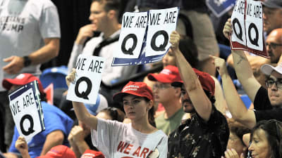 Vaalitilaisuuden yleisössä joukko ihmisiä pitää esillä kylttejä, joissa lukee "We are Q" eli "Me olemme Q".