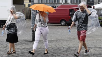 Tre personer går ute i regnet i Helsingfors. Två av dem bär en engångsregnjacka och en av dem går under ett paraply.