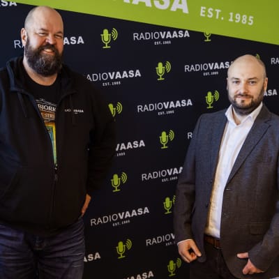 Två manspersoner står tillsammans och ser in i kameran, i bakgrunden syns Radio Vasas lokaler.