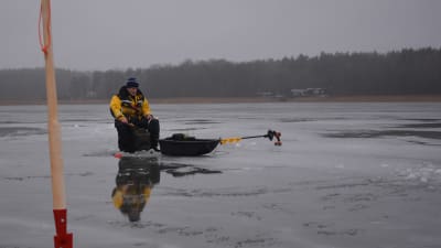 I förgrunden står en isbill med röd spets. En man i flytdräkt, en gul overall och blå mössa, sitter på en ryggsäck med pall och pimpelfiskar på en sjö där mycket smältvatten samlats på isen.
