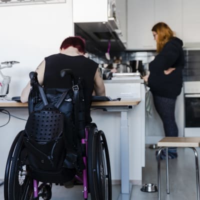 Kuvassa pyörätuolissa istuva nainen on etualalla selin kameraan kirjoituspöydän ääressä. Taustalla keittiö, jossa hääräilee avustaja. 