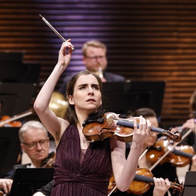 Armenialainen viulisti Diana Adamyan soittaa orkesterin edessä katse suunnattuna yläviistoon ja jousikäsi kohotettuna.