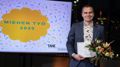 Filip Saxén står iklädd kostym och håller i ett diplom och en blombukett. Till vänster syns en teveskärm där det står "Miehen työ 2020".