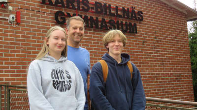 Två ungdomar (en flicka, en pojke) och bakom en man. De står ute framför en röd tegelbyggnad. På väggen står Karis-Billnäs gymnasium