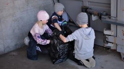 Tre barn plockar skräp i en stor sopsäck
