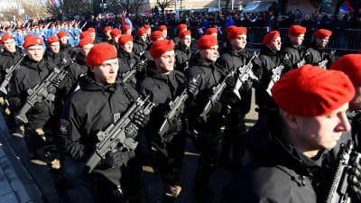 Män i svarta uniformer och röd basker marscherar i räta led med kpistar på bröstet.