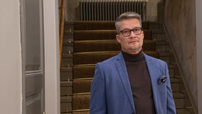En man i blå kavaj står framför en trappa i ett äldre hus. Han tittar in i kameran.