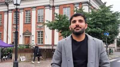 En man med mörkt hår, mustasch och skägg, svart t-skjorta och en yllekappa står på ett lite torg, med träd och en stor tegelbyggnad i bakgrunden.