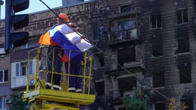 Två personer i en skylift fäster en rysk flagga i en vajer som är spänd över en gata. Bakom dem syns en skadad byggnad med utbrunna fönster.