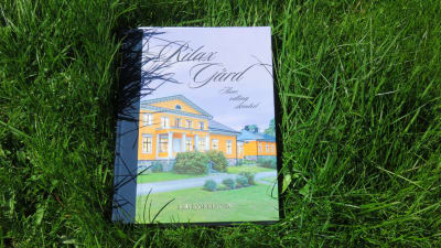 En bok som ligger på en grön oklippt gräsmatta. Pärmen visar en bild på ett stort gult hus. Boken heter Rilax gård Anor, odling, skönhet.