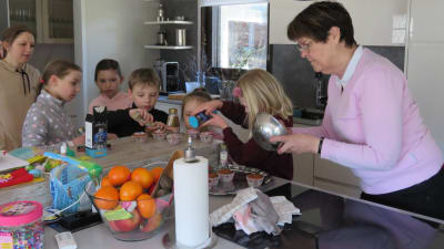Barn dekorerar muffins, cupcakes i ett kök. En kvinna ser på och en annan lagar glasyr och hjälper barnen.