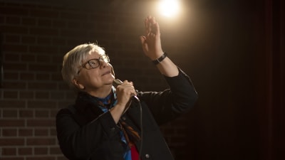 Tiina Henkola vetää stan up-keikkaa ja katsoo ylös vasen käsi ylhäällä ja oikeassa kädessä mikki.