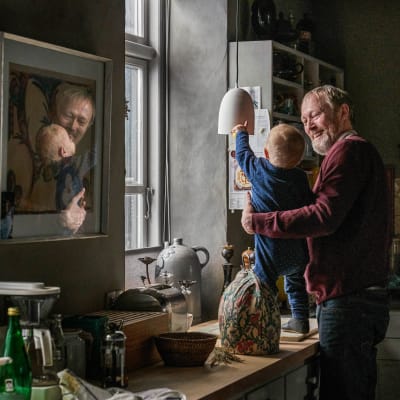 Karaktären Johannes står i ett mörkt kök och tittar skrattade på sitt barnbarn Anton.