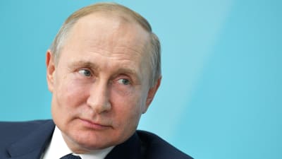 Närbild av Vladimir Putin som tittar åt sidan medan han böjer sig framåt något. Turkos bakgrund.