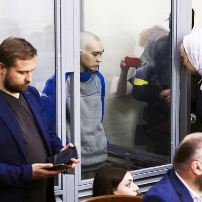  Vadim Shishimarin lasikopissa. Ympärillä ihmisiä, huivipäinen henkilö nojaa kopin ulkopuolella päätään karmia vasten.